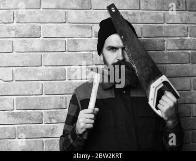 bearded worker man, long beard, brutal caucasian hipster with moustache in black cap holding various building tools: saw and hammer with serious face on brick wall studio background Stock Photo