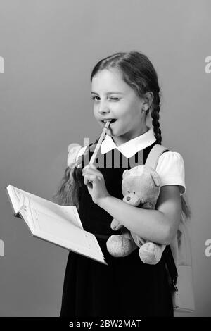 Back to school and education concept. Girl holds teddy bear, open notebook and green marker. School girl with flirty smile isolated on green background. Pupil in school uniform with braids and bag Stock Photo