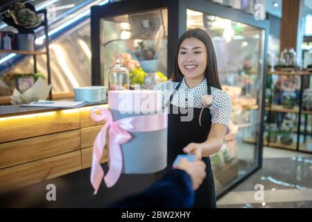 Brunette female in apron giving round gift box with pink bow to customer, receiving credit card from him Stock Photo