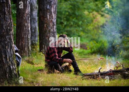 Guy with tired face and lonely at picnic or barbecue. Man, hipster, hiker roast sausages on stick on bonfire in forest. Summer camping, hiking, vacation concept. Hipster with beard cooking food. Stock Photo