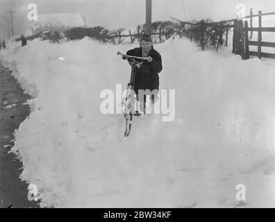 Drifts of snow four feet deep block Surrey roads to traffic . Drifts of snow more than four feet in depth have blocked roads in Surrey and Kent and made them impassable to traffic , armies of men are at work clearing the roads . A young boy fights on his bike through the snow . 13 February 1932 Stock Photo