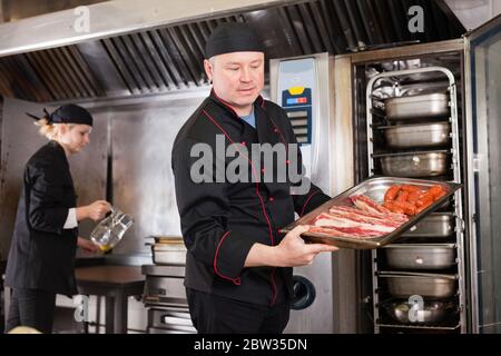 Skilled cook working in professional kitchen, preparing to put pork ribs and sausages into oven Stock Photo