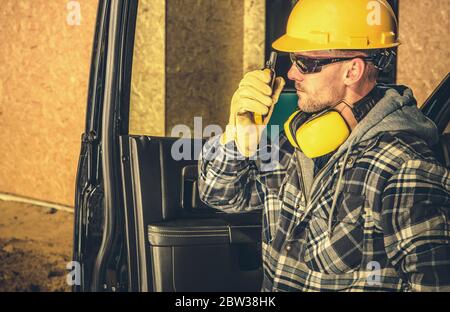 Caucasian Contractor Making Quick on Site Conversation with Other Workers Using Walkie Talkie While Staying Side to His Business Vehicle. Construction Stock Photo