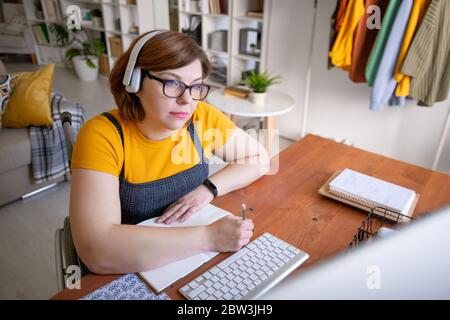 Focused young woman in wireless headphones sitting in wheelchair and making notes about lecture while studying at home Stock Photo