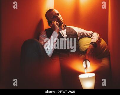 A mature handsome caucasian man entrepreneur in a checkered vest is sitting on a cushion in a cozy nook of a coworking and talking on a cellphone Stock Photo
