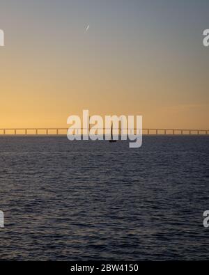A single sailing boat is out on the the calm ocean between Denmark and Sweden during sunset with the Oresund bridge in the background. Stock Photo