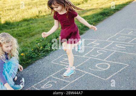 Cute adorable children girls friends playing jumping hopscotch outdoor. Funny activity game for kids on playground outside. Summer backyard street Stock Photo
