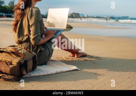 woman blogger working remotely with a laptop using the internet, on the seashore, sitting on the beach, Holds a laptop on her lap. Work on vacation wh Stock Photo