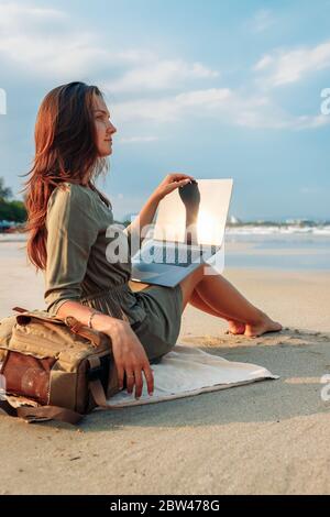 Young woman blogger working remotely with a laptop using the internet, on the seashore, sitting on the beach. Work on vacation while traveling. Freela Stock Photo