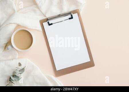 Home office desk with blank clipboard, cup of coffee, blanket and eucalyptus leaf. Modern feminine workspace, minimal style. Flat lay, top view. Stock Photo