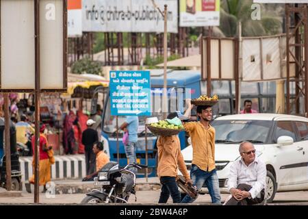 Somnath, Gujarat, India - December 2018: Two street vendors smiling and walking on a crowded market street carrying a basket of fruits on their heads. Stock Photo