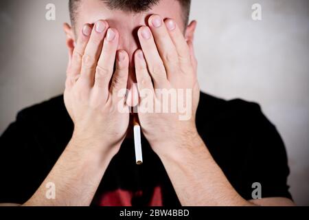 man with a cigarette in his mouth shyly covers his face with his palms, hiding his eyes Stock Photo