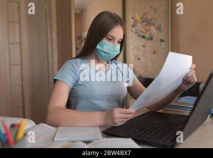 Teenager girl in protective medical mask, in hands letter of test admission leaflet, security viral infections, home electronic education, laptop and Stock Photo