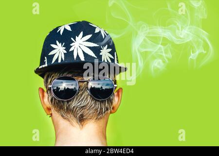 Girl in sunglasses and cap with leaves of marijuana smokes on green background Stock Photo