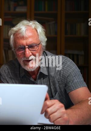 Portrait of a white bearded senior man with grey shirt smiling while looking at a laptop, notebook screen. Stock Photo