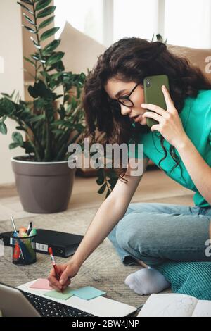 Busy caucasian woman with curly hair making notes sitting at the computer and having a phone discussion Stock Photo