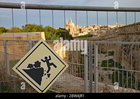 Sign attached to metal fence at Fort Manoel in Malta warning of the dangers of falling. The dome of St Paul's church in Valletta can be seen in the ba Stock Photo