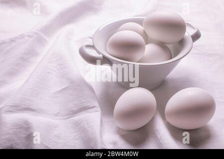on the white linen tablecloth, in the foreground, a ceramic bowl with fresh white eggs ready for the preparation of homemade culinary recipes Stock Photo