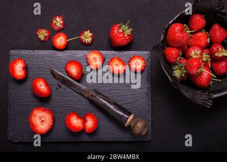 seen from above. Bowl with strawberries, on a slate stone background some red strawberries sectioned with an old knife Stock Photo