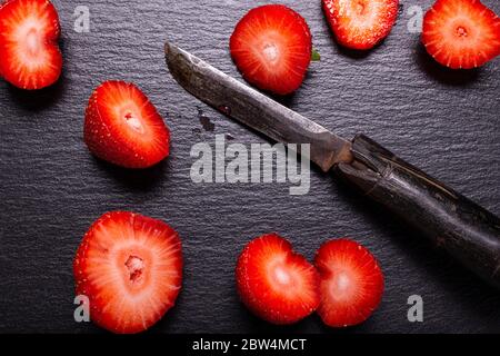 seen from above. On a slate stone background some red strawberries sectioned with an old knife Stock Photo