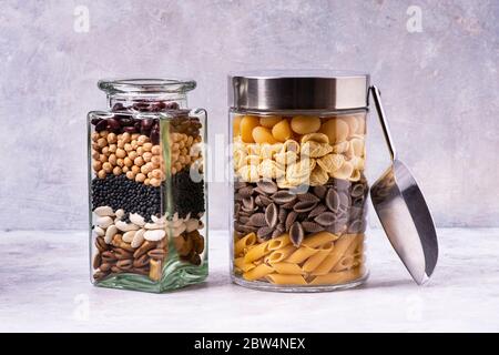 On a gray textured background, varieties of Italian pasta and dried legumes in glass jars in the foreground. Mediterranean diet Stock Photo