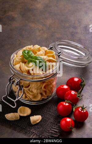 in the foreground in a transparent glass jar of raw Italian pasta and fresh basil leaves. in the background bunch of cherry tomatoes Stock Photo