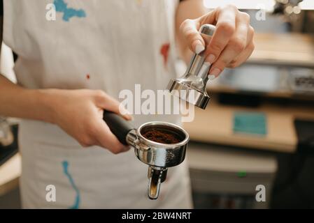 A female barista holds a metal tamper and a portafilter with coffee in a coffee shop. A barista preparing for pressing ground coffee for brewing espre Stock Photo