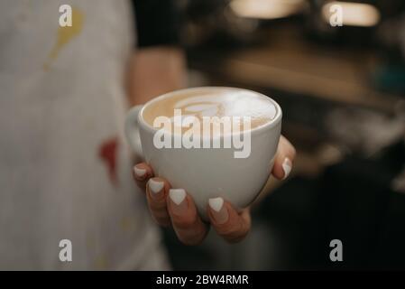 A female barista holds a cup of hot latte in her hand in a cafe. A waitress preparing a customer order. Stock Photo