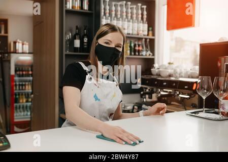 A female barista in a medical black face mask smiles and cleaning the bar counter in a coffee shop. A beautiful waitress disinfecting a cafe. Stock Photo