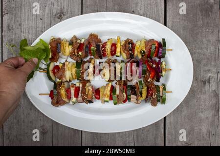 Skewer before grilling composed of bacon onions tomatoes meat pork zucchini peppers on a plate with leaves and hand on a wooden table Stock Photo