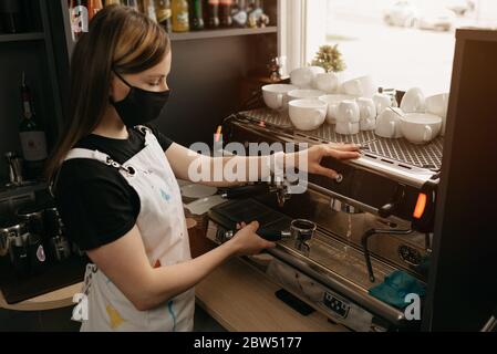 A happy female barista in a medical black face mask works with a coffee machine in the coffee shop. Stock Photo