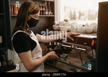 A happy female barista in a medical black face mask works with a coffee machine in the coffee shop. Stock Photo