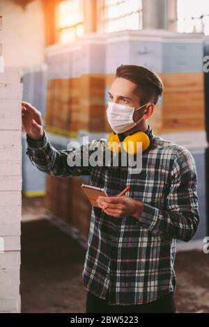 Professional man supervisor in medical face mask and protective headphones inspecting wood stock inventory. Male manager, warehouse worker checking, c Stock Photo