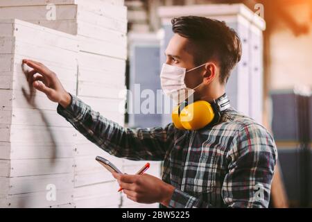 Portrait male foreman inspector in medical face mask and protective headphones checking woodwork stock at factory storage. Man supervisor counting woo Stock Photo