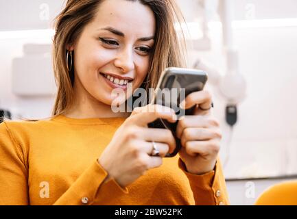 Happy woman smiling and browsing social media on smartphone while visiting contemporary dental clinic Stock Photo