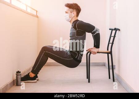 Young confident sports man in medical face mask doing triceps dip on chair at home. Sporty athletic man fitness instructor in protective mask exercisi Stock Photo