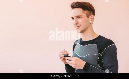 Portrait young confident sports man listening music with wireless earbuds and smiling. Happy fitness man in bluetooth earphones against background wit Stock Photo