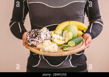 Closeup young sports man hold in hands wooden board with fresh apples, bananas, vegetables, donut and chocolate. Fitness man struggling for choice bet