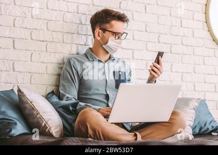 Young businessman in medical face mask using smart phone, texting message while working with laptop at home office. Happy hipster man hold mobile phon Stock Photo