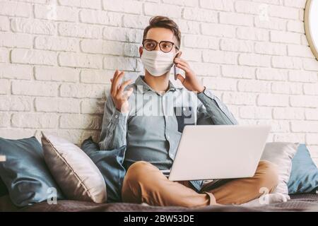 Happy hipster man freelancer in medical face mask talking mobile phone, sitting on couch with laptop on laps. Young businessman in protective mask wor Stock Photo