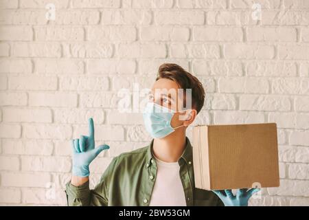 Young hipster guy delivery service courier in medical face mask hold hold cardboard box in hand look up and point finger at copy space. Happy delivery Stock Photo