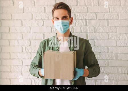 Hipster man courier in medical face mask and rubber gloves hold cardboard box in hands. Happy man delivery service employee in protective face mask ca Stock Photo