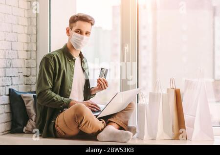 Happy man shopper in medical face mask hold credit card in hand for purchases in internet store. Young hipster man in protective mask using laptop for Stock Photo