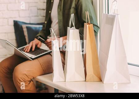 Closeup young hipster man using laptop for online shopping and holding credit card in hand, sitting on windowsill with bags. Stylish man shopper buyin Stock Photo