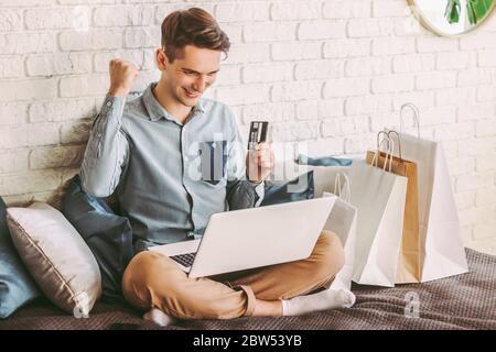 Happy male shopper shopping online with laptop, hold credit card in hand and smiling on couch. Young stylish hipster man celebrating successful purcha Stock Photo