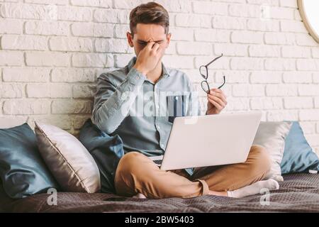 Young tired businessman feeling headache while sitting on couch with laptop on laps. Exhausted stressed hipster man freelancer blogger feel frustrated Stock Photo