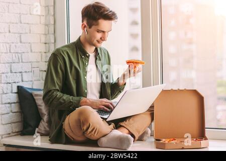 Young hipster man freelancer eating slice of tasty pizza, sit on windowsill and using laptop for study, distance learning. Businessman eat takeaway it Stock Photo
