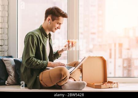 Happy businessman eat slice of tasty pizza, work remotely from home. Young hipster man freelancer sit on windowsill, using laptop for distance educati Stock Photo
