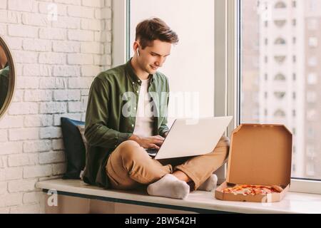 Happy businessman using laptop for distance work from home and eat tasty takeaway pizza fast food. Young hipster man freelancer typing on computer key Stock Photo