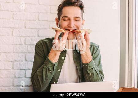 Hungry hipster man freelancer eating tasty italian pizza while studying remotely from home. Happy young businessman hold takeaway fast food pizza slic Stock Photo